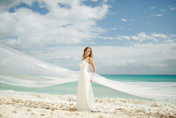 Невеста стоит на пляже в белом, длинном платье, держась за прозрачную ткань, на фоне синего неба - фото 1898987 Travel Service Mexico - свадьба в Мексике