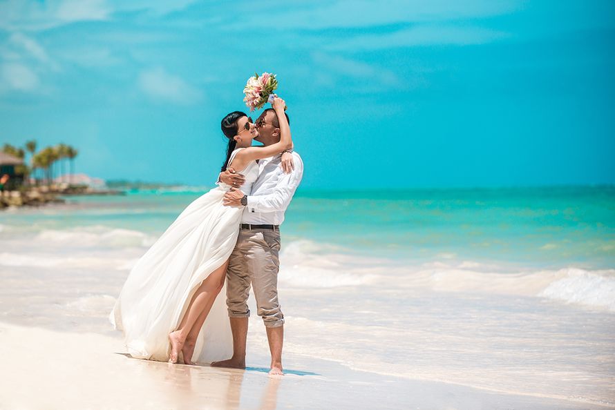 В лазурном прибое парень в белой рубашке и бежевых брюках, обняв за талию, целует невесту в белоснежном длинном платье с глубоким - фото 2939477 Свадебное агенство "Travelservice weddings"
