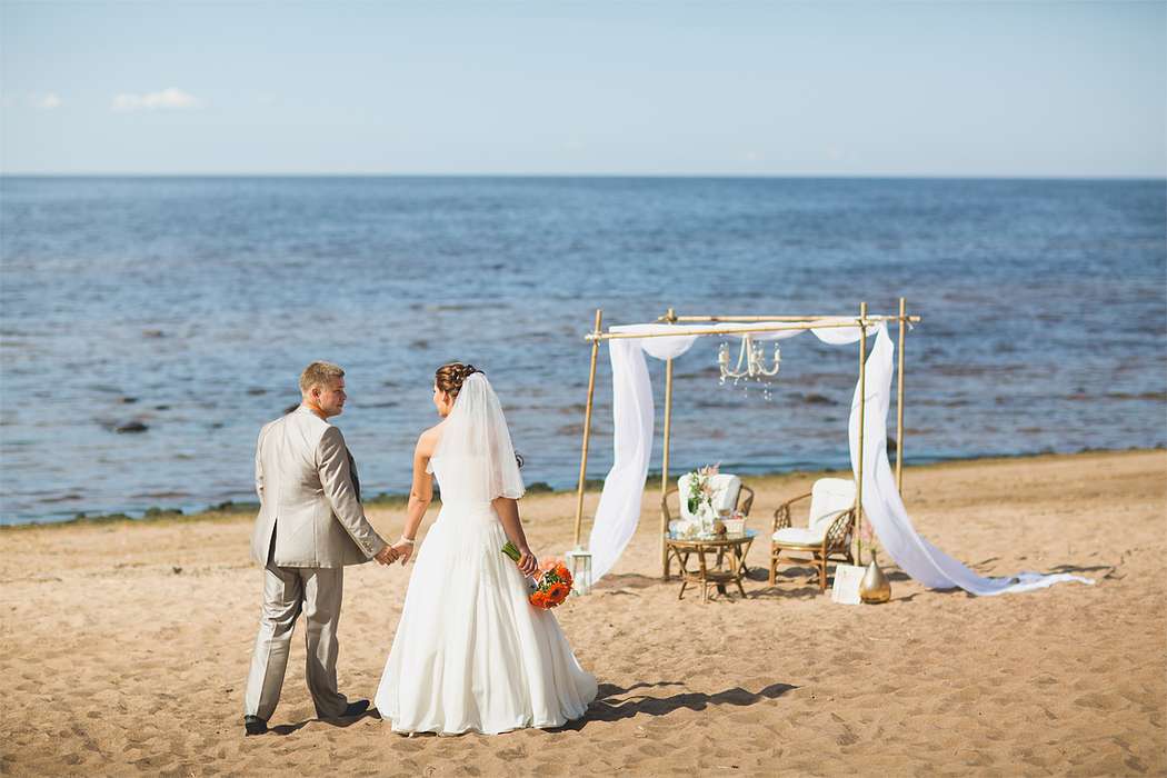 На побережье стоит арка из бамбука в белых тканях, на ней обручальные кольца, стол из лозы и два стула, по пляжу идут жених и - фото 2037294 Студия свадеб и декора "Мариаж"