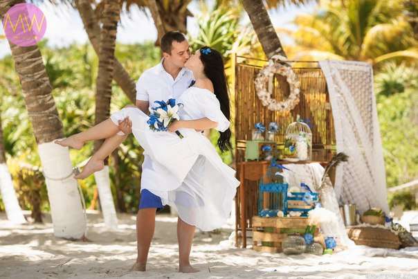 По тропическому побережью жених в синих шортах и белой рубашке несет на руках невесту в открытом белом платье, целуя ее, у нее в - фото 2369476 Mainland Travel Bureau - свадьба за границей