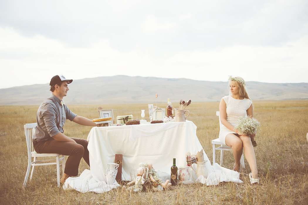 Жених и невеста сидят посреди поля за маленьким столиком, украшенным белой скатертью - фото 1596501 Фотограф Манвелов Николай