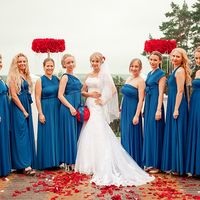 На полу с красными лепестками стоит невеста с красными цветами в волосах, фате, в платье со шлейфом и кружевом, с букетом-шариком на руке из красных роз декорированный синей лентой  и подружки в длинных синих платьях трасформер