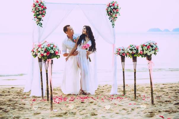 На пляже под белой аркой стоят жених с невестой, он оперся на белый столик и смотрит на нее, в белой рубашке, она в длинном - фото 2832725 Romantica - свадебное агентство в Таиланде