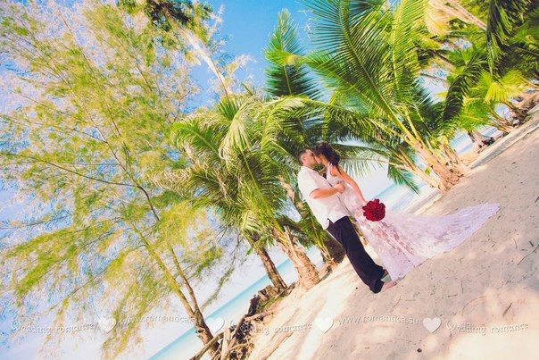 На песчаном побережье в тени тропических деревьев целуются молодожены, жених в черных брюках и белой рубашке держит за талию - фото 2832731 Romantica - свадебное агентство в Таиланде