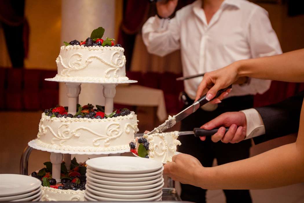 Жених с невестой, разрезающий  двухъярусный белый 
свадебный торт, украшенный ягодами и фруктами  - фото 1213589 Люсьена Солнечная